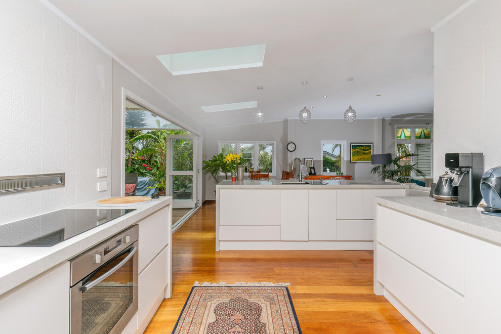 Inspiration for a modern galley kitchen/diner in Auckland with a double-bowl sink, white cabinets, composite countertops, white splashback, porcelain splashback, stainless steel appliances, medium hardwood flooring, an island and white worktops.
