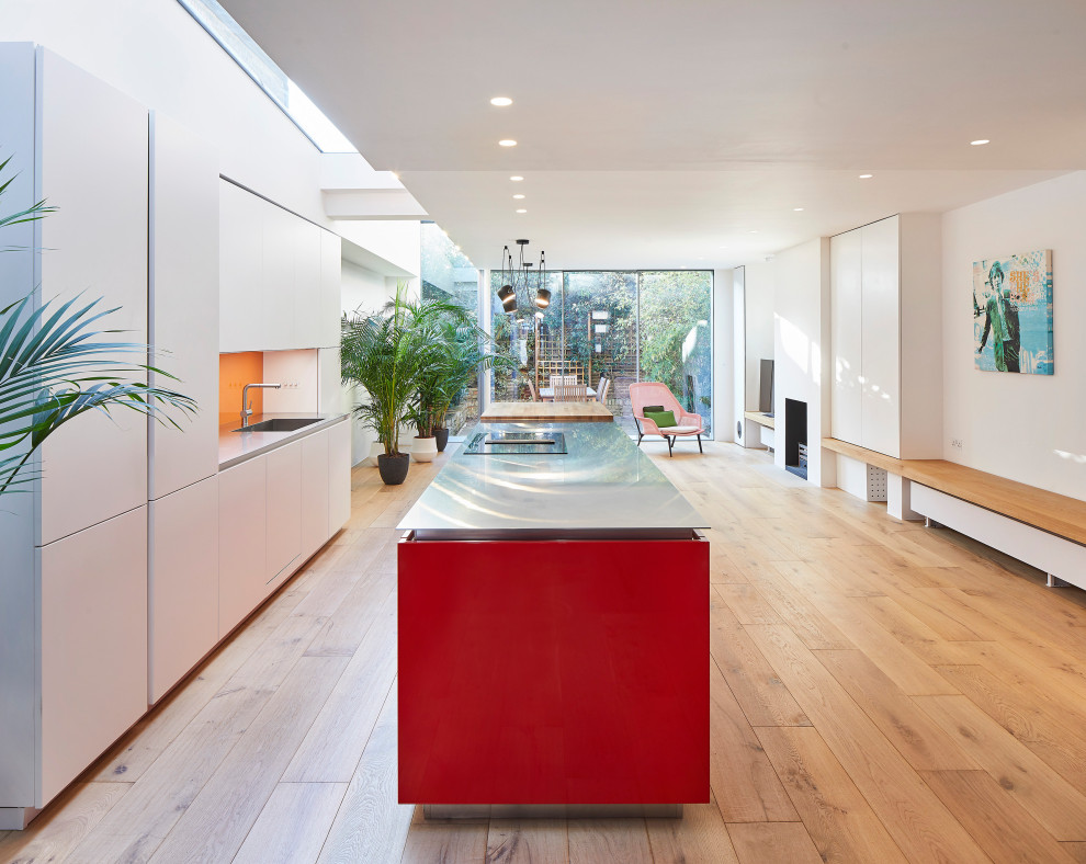 Large contemporary open plan kitchen in London with an integrated sink, flat-panel cabinets, red cabinets, stainless steel worktops, orange splashback, glass sheet splashback, integrated appliances, light hardwood flooring, an island, beige floors and grey worktops.