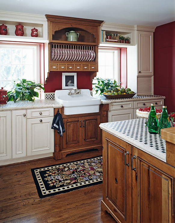 Landhausstil Wohnküche in L-Form mit Landhausspüle, profilierten Schrankfronten, Arbeitsplatte aus Fliesen, Küchenrückwand in Rot, braunem Holzboden, Kücheninsel und Elektrogeräten mit Frontblende in Sonstige