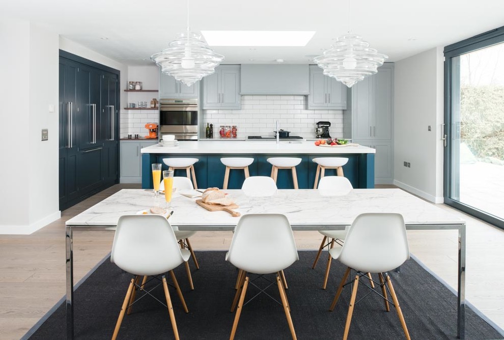 Inspiration for a large contemporary single-wall open plan kitchen in Kent with a built-in sink, recessed-panel cabinets, blue cabinets, composite countertops, white splashback, ceramic splashback, stainless steel appliances, light hardwood flooring and an island.