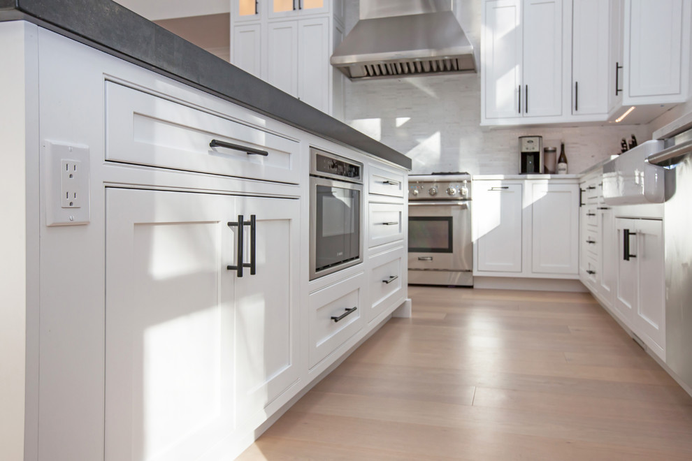 Photo of a large modern l-shaped kitchen/diner in Boston with a belfast sink, beaded cabinets, white cabinets, granite worktops, white splashback, glass tiled splashback, stainless steel appliances, medium hardwood flooring, an island and brown floors.