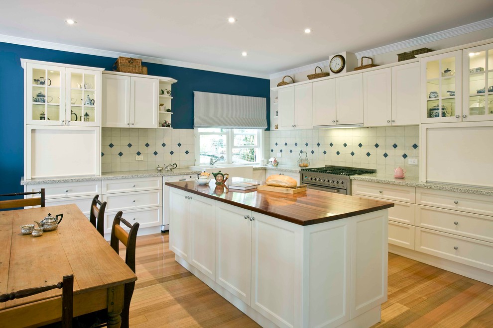 Klassisk inredning av ett stort kök, med vita skåp, luckor med infälld panel, en rustik diskho, träbänkskiva, vitt stänkskydd, stänkskydd i terrakottakakel, rostfria vitvaror, ljust trägolv och en köksö