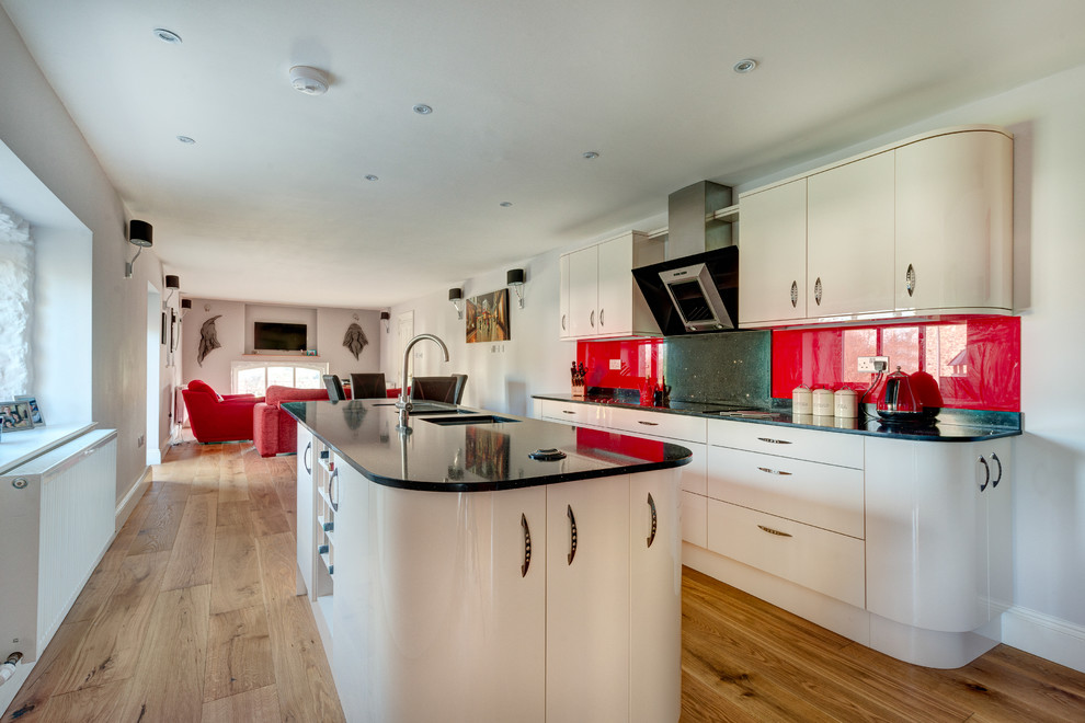 Offene, Zweizeilige, Mittelgroße Moderne Küche mit Unterbauwaschbecken, flächenbündigen Schrankfronten, Granit-Arbeitsplatte, Küchenrückwand in Rot, Glasrückwand, Küchengeräten aus Edelstahl, braunem Holzboden und Kücheninsel in Devon