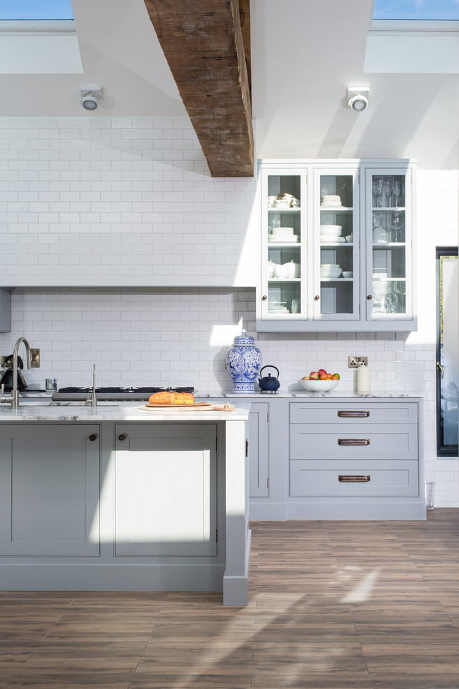 Example of a large arts and crafts single-wall dark wood floor open concept kitchen design in Dublin with a farmhouse sink, glass-front cabinets, gray cabinets, marble countertops, white backsplash, ceramic backsplash, stainless steel appliances and an island