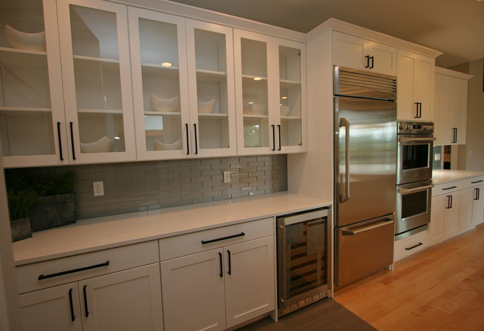 Bild på ett mellanstort funkis kök, med en undermonterad diskho, skåp i shakerstil, vita skåp, bänkskiva i kvarts, grått stänkskydd, stänkskydd i glaskakel, rostfria vitvaror, ljust trägolv och en köksö