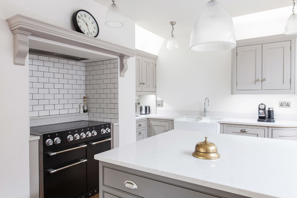 This is an example of a medium sized classic l-shaped kitchen/diner in Kent with a belfast sink, beaded cabinets, grey cabinets, quartz worktops, white splashback, metro tiled splashback, black appliances, light hardwood flooring, an island and beige floors.