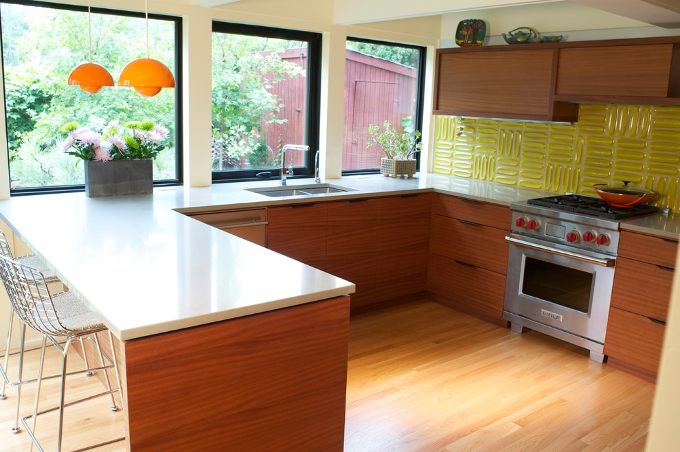 Modern inredning av ett mellanstort u-kök, med en dubbel diskho, släta luckor, skåp i mellenmörkt trä, bänkskiva i kvarts, gult stänkskydd, rostfria vitvaror, ljust trägolv och en halv köksö