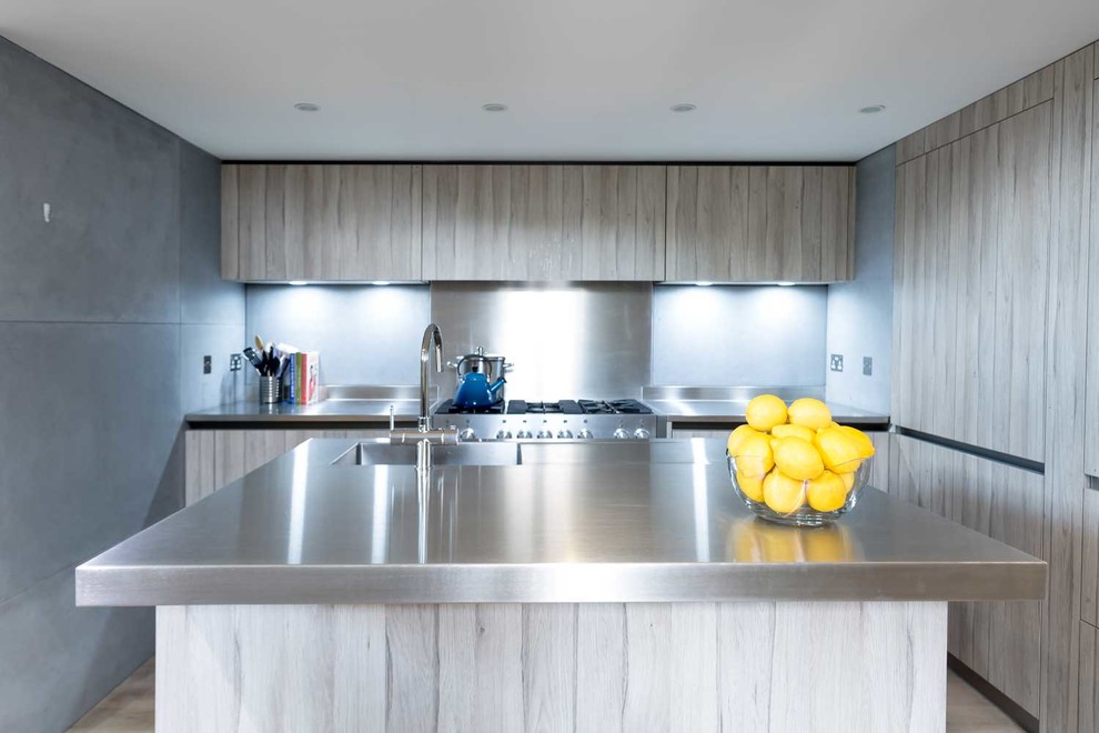 Photo of a medium sized contemporary l-shaped open plan kitchen in London with an integrated sink, flat-panel cabinets, light wood cabinets, stainless steel worktops, grey splashback, cement tile splashback, stainless steel appliances, light hardwood flooring and an island.