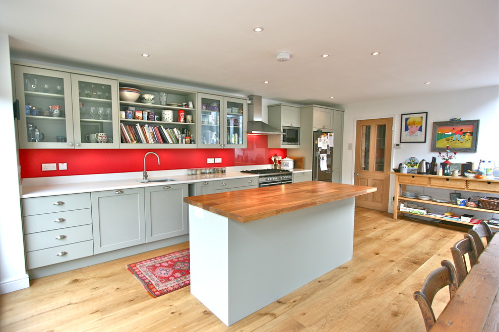 Design ideas for a medium sized contemporary single-wall kitchen/diner in London with a submerged sink, shaker cabinets, green cabinets, engineered stone countertops, red splashback, glass sheet splashback, stainless steel appliances, light hardwood flooring and an island.