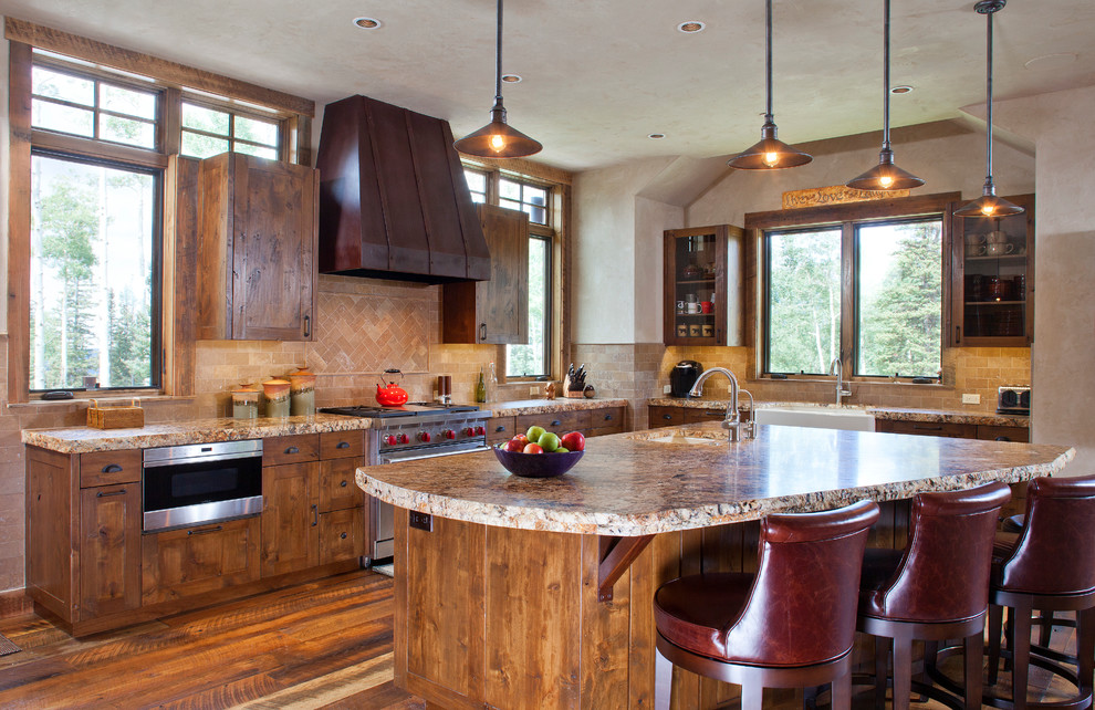 Cette image montre une cuisine chalet en L et bois brun avec îlot, un évier de ferme, un placard à porte shaker, une crédence beige, un électroménager en acier inoxydable et un sol en bois brun.