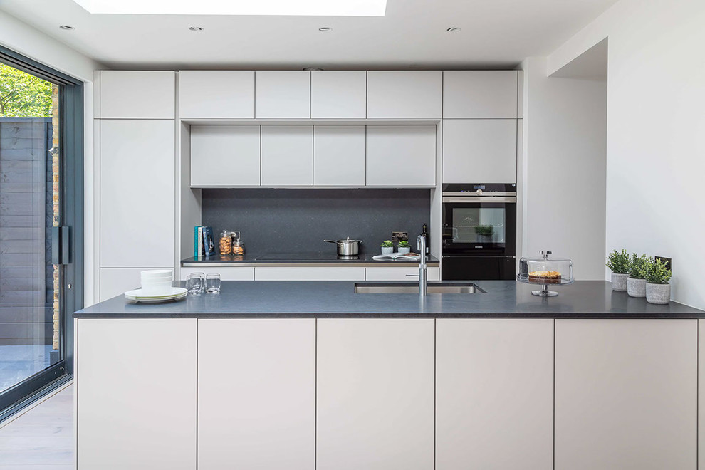 Contemporary kitchen in Hertfordshire with a submerged sink, flat-panel cabinets, grey cabinets, grey splashback, black appliances, a breakfast bar and grey worktops.