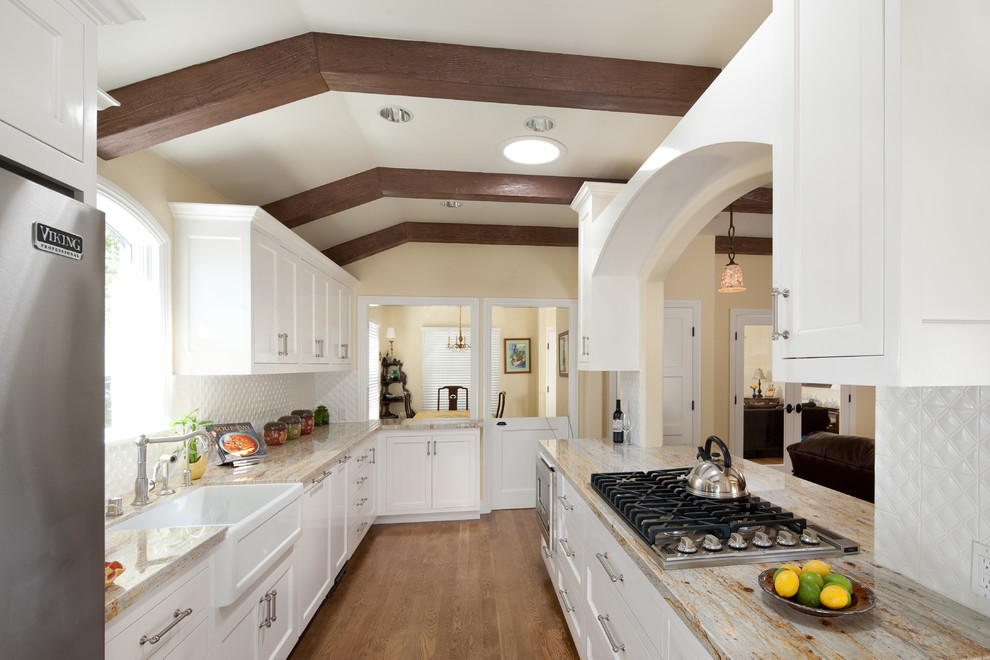 Eat-in kitchen - mid-sized traditional galley medium tone wood floor eat-in kitchen idea in Los Angeles with a farmhouse sink, shaker cabinets, white cabinets, white backsplash, glass tile backsplash, stainless steel appliances and a peninsula