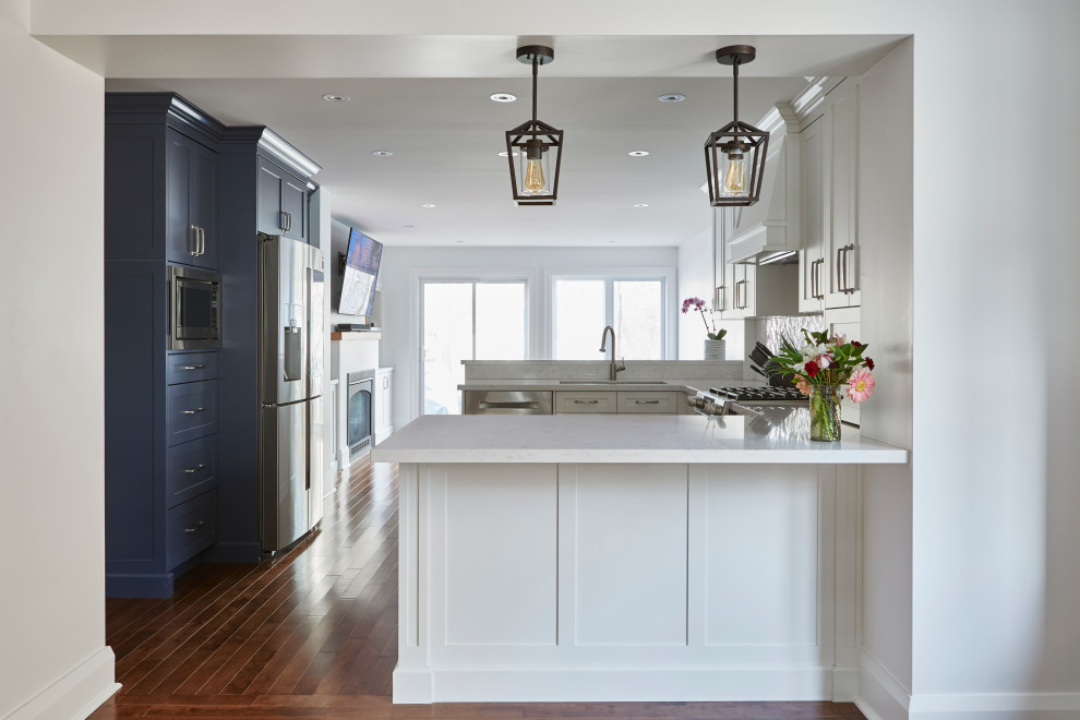 Modern inredning av ett mellanstort vit vitt kök, med en undermonterad diskho, skåp i shakerstil, vita skåp, bänkskiva i kvarts, vitt stänkskydd, stänkskydd i keramik, rostfria vitvaror, mellanmörkt trägolv, en halv köksö och brunt golv