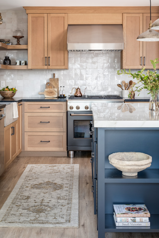 Example of a mid-sized transitional l-shaped vinyl floor and beige floor kitchen design in San Diego with a farmhouse sink, shaker cabinets, blue cabinets, solid surface countertops, white backsplash, terra-cotta backsplash, black appliances, an island and black countertops