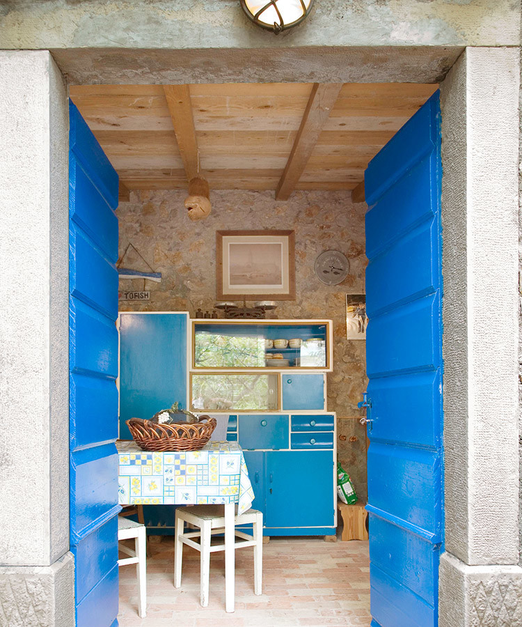Medelhavsstil inredning av ett litet kök, med en enkel diskho, öppna hyllor, blå skåp, träbänkskiva, beige stänkskydd, stänkskydd i sten, vita vitvaror och tegelgolv