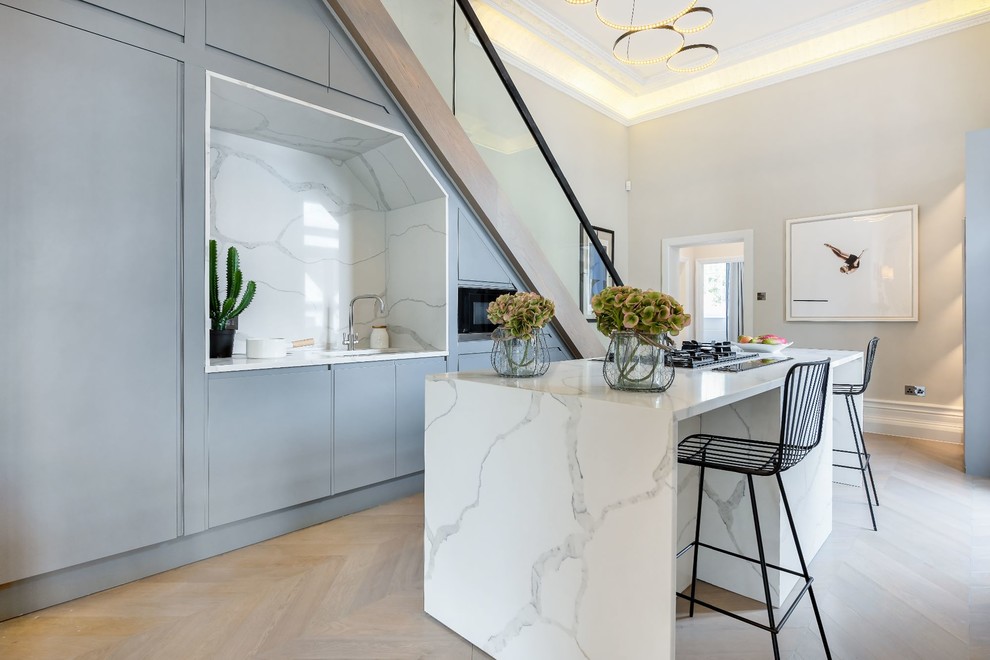 Inredning av ett modernt vit vitt kök med öppen planlösning, med en enkel diskho, släta luckor, grå skåp, marmorbänkskiva, vitt stänkskydd, stänkskydd i marmor, ljust trägolv, en köksö och beiget golv