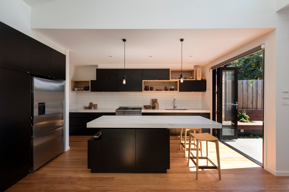 Contemporary open plan kitchen in Melbourne with a submerged sink, flat-panel cabinets, black cabinets, marble worktops, white splashback, metro tiled splashback, stainless steel appliances, light hardwood flooring and an island.
