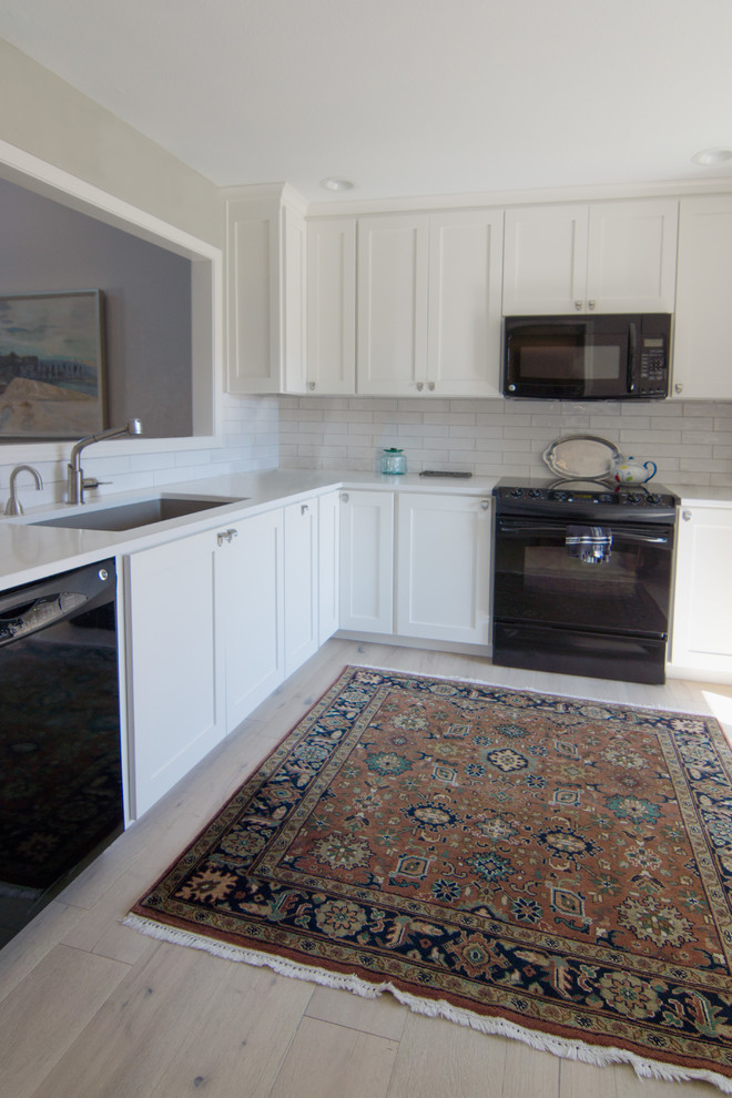 This is an example of a bohemian l-shaped kitchen in Kansas City with recessed-panel cabinets, white cabinets, white splashback, porcelain splashback, black appliances, light hardwood flooring, no island and white worktops.