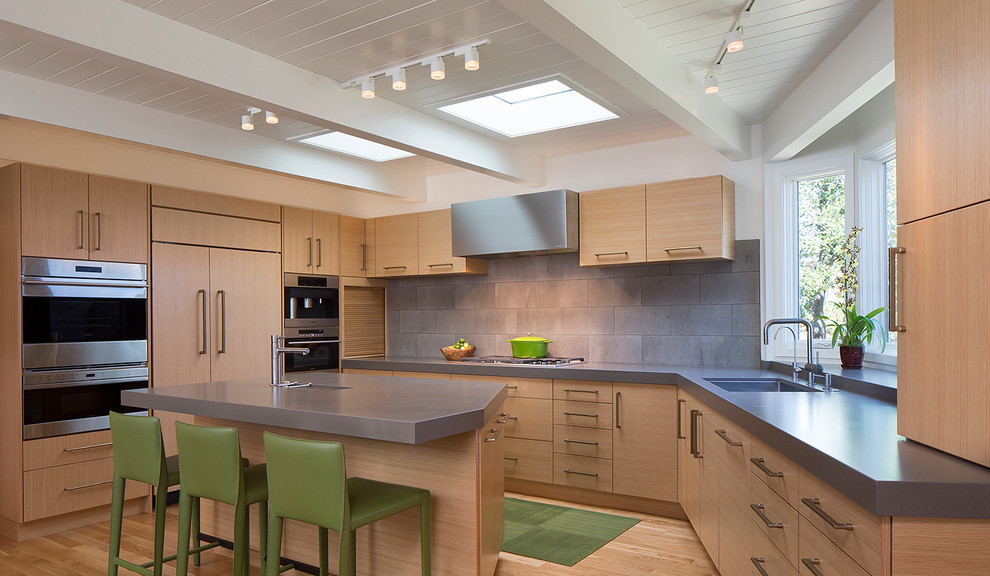 Foto på ett funkis kök, med en undermonterad diskho, släta luckor, skåp i ljust trä, grått stänkskydd, rostfria vitvaror, ljust trägolv och en köksö
