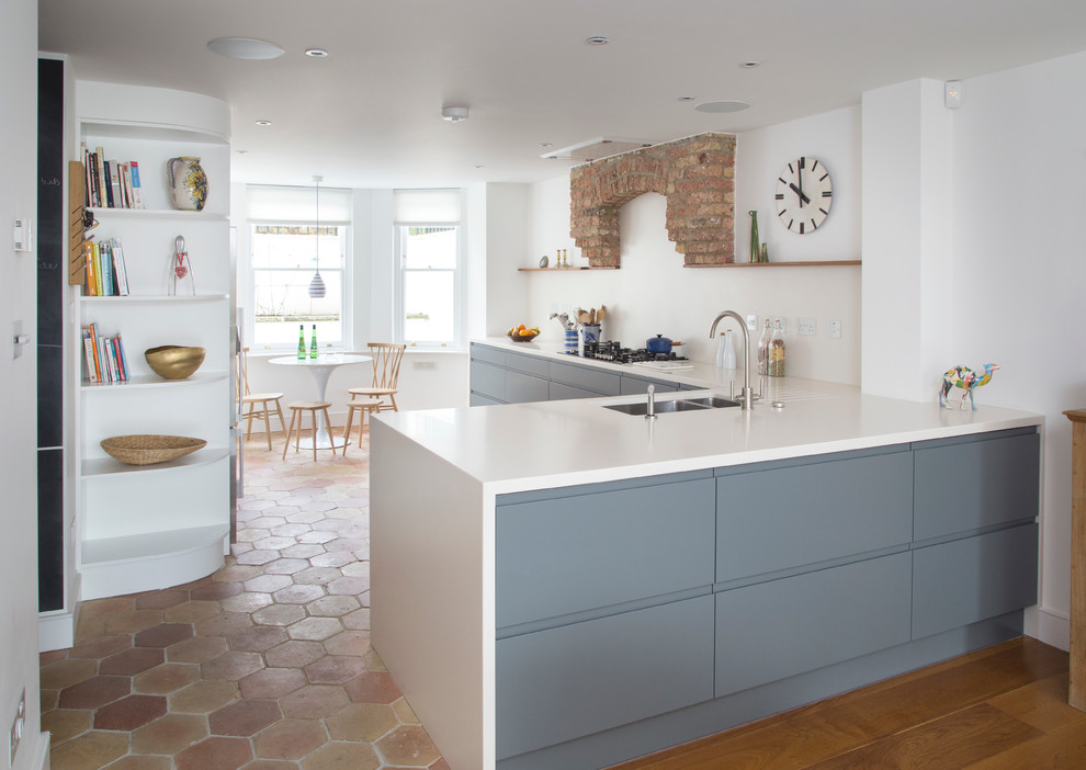 Design ideas for a large contemporary l-shaped kitchen/diner in London with a double-bowl sink, flat-panel cabinets, blue cabinets, composite countertops, white splashback, stainless steel appliances, terracotta flooring, an island, multi-coloured floors, white worktops and a feature wall.
