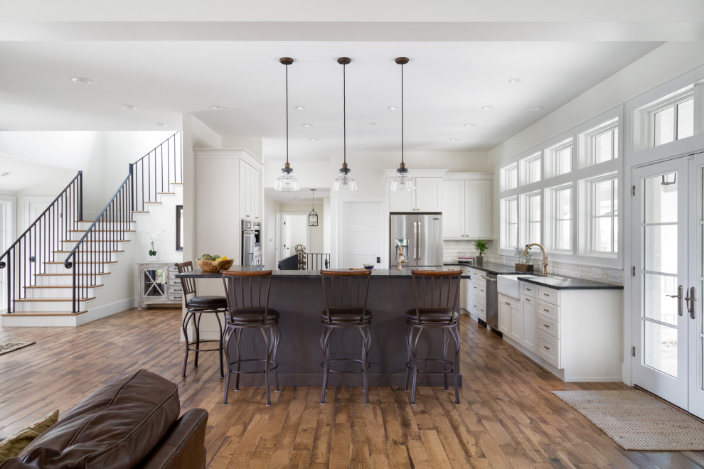 Inspiration for a large transitional u-shaped medium tone wood floor and brown floor open concept kitchen remodel in Denver with a farmhouse sink, shaker cabinets, white cabinets, white backsplash, stainless steel appliances, an island and black countertops
