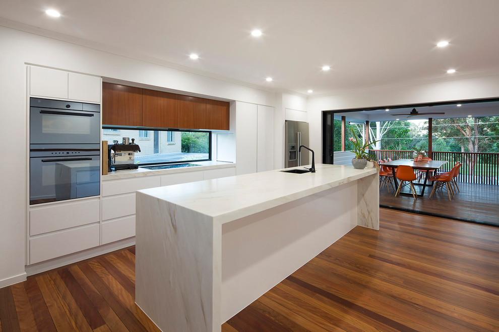 Design ideas for a medium sized modern galley open plan kitchen in Brisbane with a submerged sink, flat-panel cabinets, white cabinets, marble worktops, glass sheet splashback, stainless steel appliances, light hardwood flooring, an island, multi-coloured floors and white worktops.