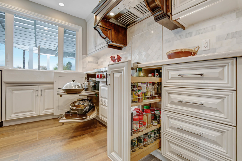 Example of a mid-sized classic l-shaped porcelain tile and brown floor open concept kitchen design in Los Angeles with a farmhouse sink, raised-panel cabinets, white cabinets, quartz countertops, white backsplash, marble backsplash, stainless steel appliances, an island and white countertops