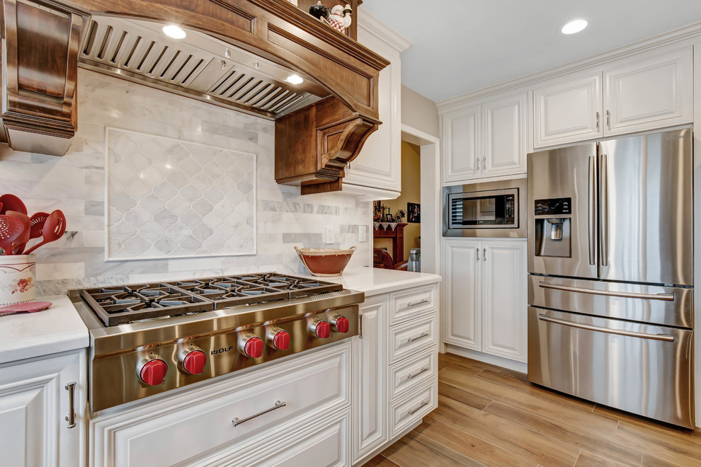 Klassisk inredning av ett mellanstort vit vitt kök, med en rustik diskho, luckor med upphöjd panel, vita skåp, bänkskiva i kvarts, vitt stänkskydd, stänkskydd i marmor, rostfria vitvaror, klinkergolv i porslin, en köksö och brunt golv