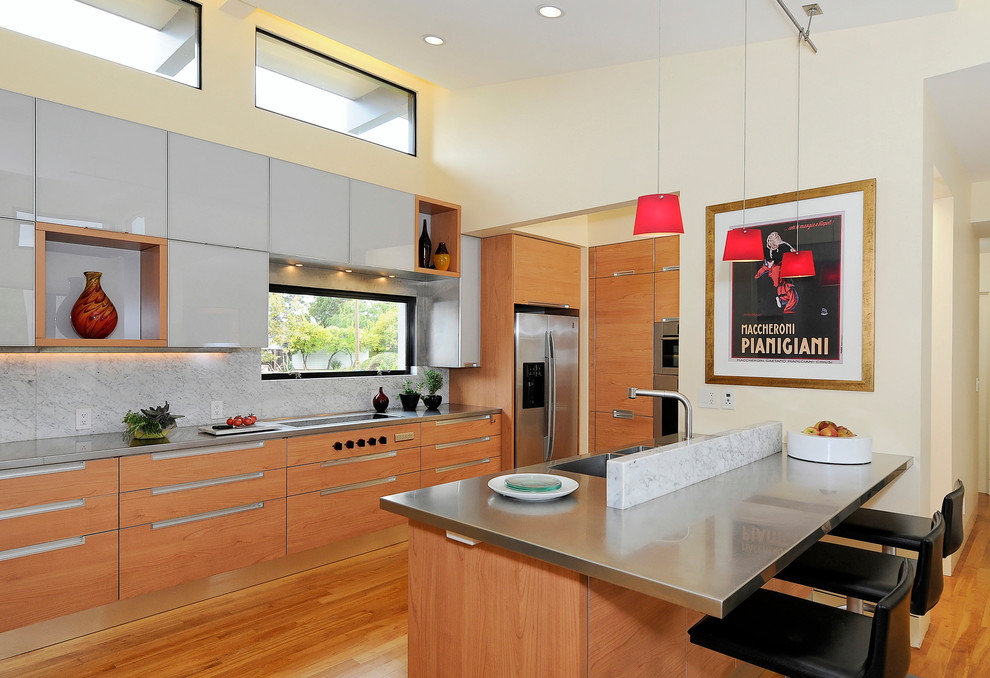 Bild på ett funkis kök, med rostfria vitvaror, bänkskiva i rostfritt stål, en integrerad diskho, släta luckor, skåp i mellenmörkt trä, vitt stänkskydd och stänkskydd i sten