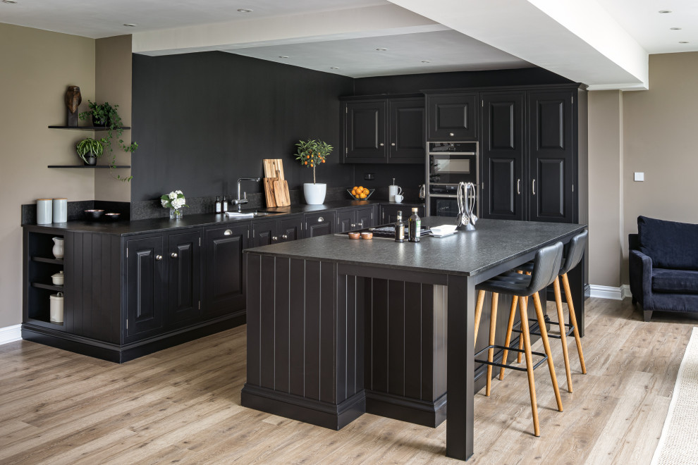 Photo of a large classic l-shaped open plan kitchen in Hampshire with raised-panel cabinets, black cabinets, an island, black worktops, a submerged sink, black appliances, medium hardwood flooring and brown floors.