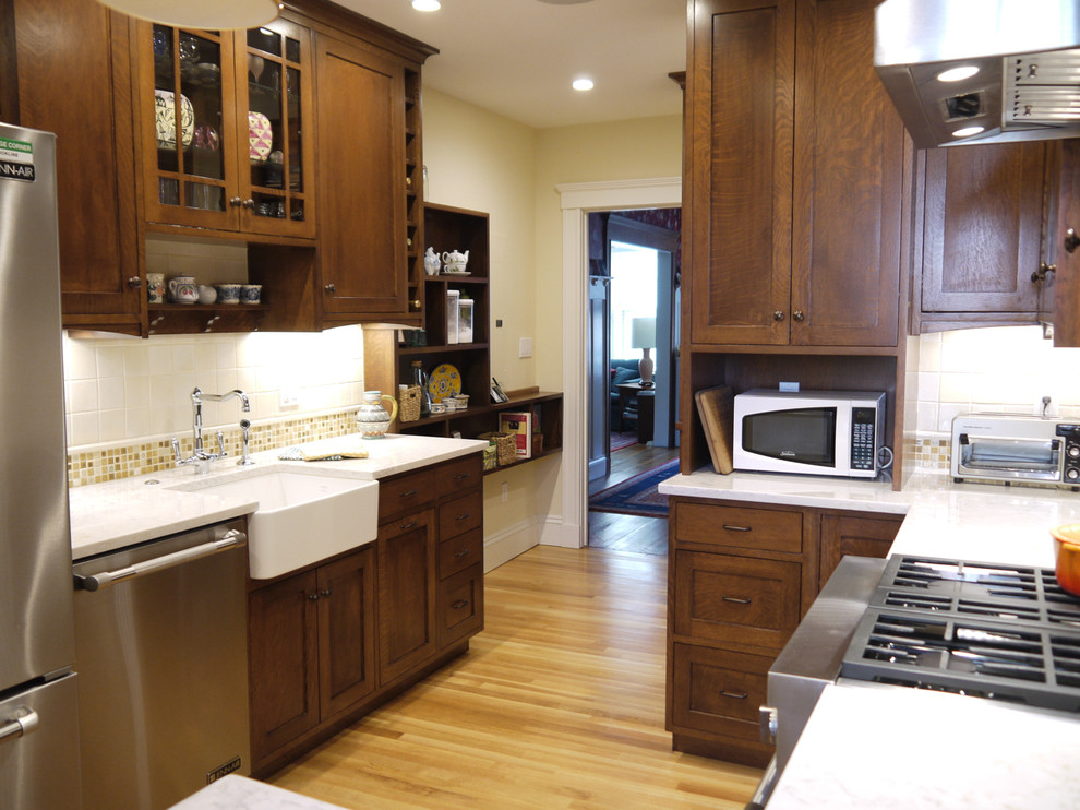 Inspiration for a mid-sized craftsman light wood floor enclosed kitchen remodel in Boston with flat-panel cabinets, medium tone wood cabinets, quartz countertops, beige backsplash, ceramic backsplash, a peninsula, a farmhouse sink and stainless steel appliances
