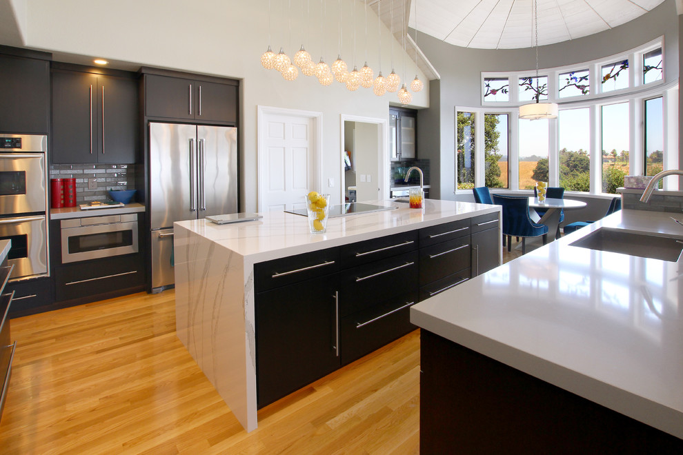 Photo of a large modern u-shaped kitchen/diner in San Francisco with a submerged sink, flat-panel cabinets, dark wood cabinets, engineered stone countertops, grey splashback, glass tiled splashback, stainless steel appliances, light hardwood flooring, an island, brown floors and white worktops.