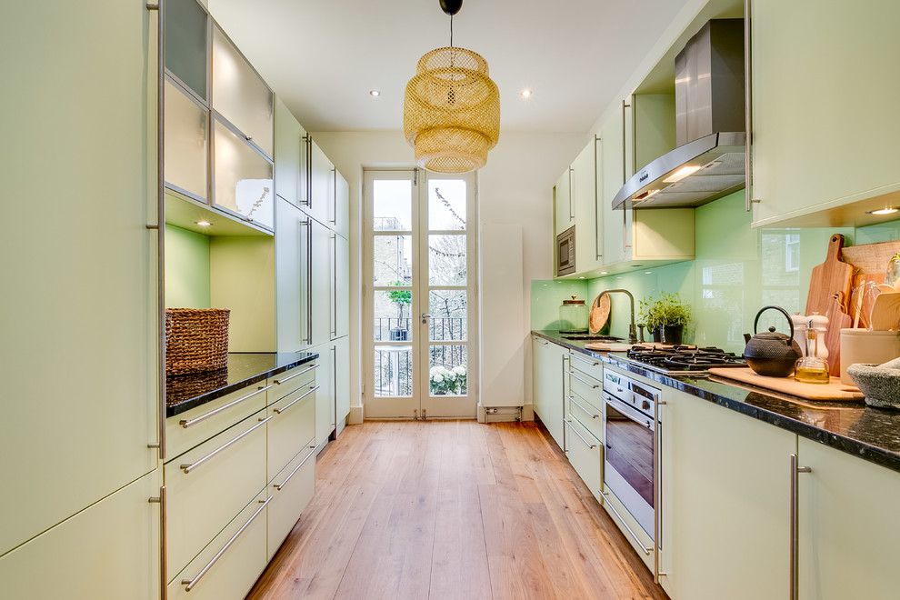 Design ideas for a contemporary galley enclosed kitchen in London with flat-panel cabinets, green cabinets, green splashback, glass sheet splashback, stainless steel appliances, light hardwood flooring, no island, brown floors and black worktops.