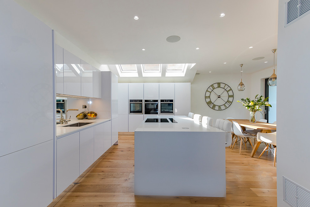 Inredning av ett modernt stort kök och matrum, med en undermonterad diskho, vita skåp, bänkskiva i kvarts, integrerade vitvaror, ljust trägolv och en köksö