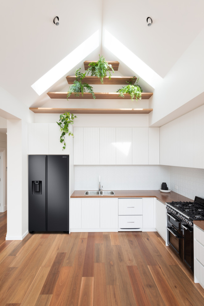 Photo of a small scandi l-shaped kitchen/diner in Melbourne with a double-bowl sink, flat-panel cabinets, white cabinets, wood worktops, white splashback, metal splashback, black appliances, medium hardwood flooring, no island, brown floors, brown worktops and a vaulted ceiling.