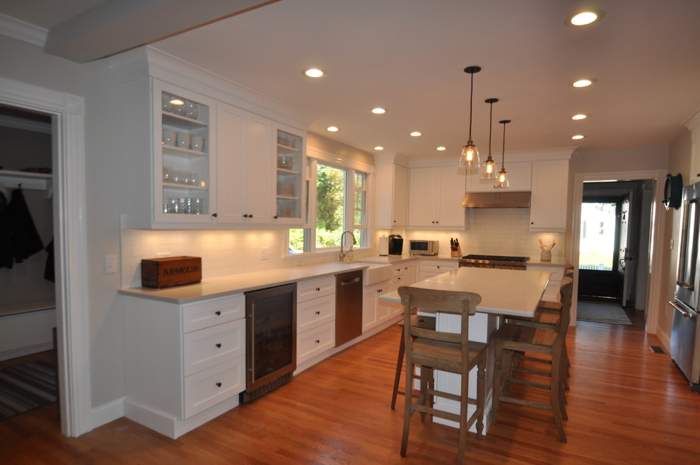 Inspiration for a mid-sized transitional l-shaped medium tone wood floor and brown floor open concept kitchen remodel in Boston with a farmhouse sink, shaker cabinets, white cabinets, quartz countertops, white backsplash, subway tile backsplash, stainless steel appliances and an island