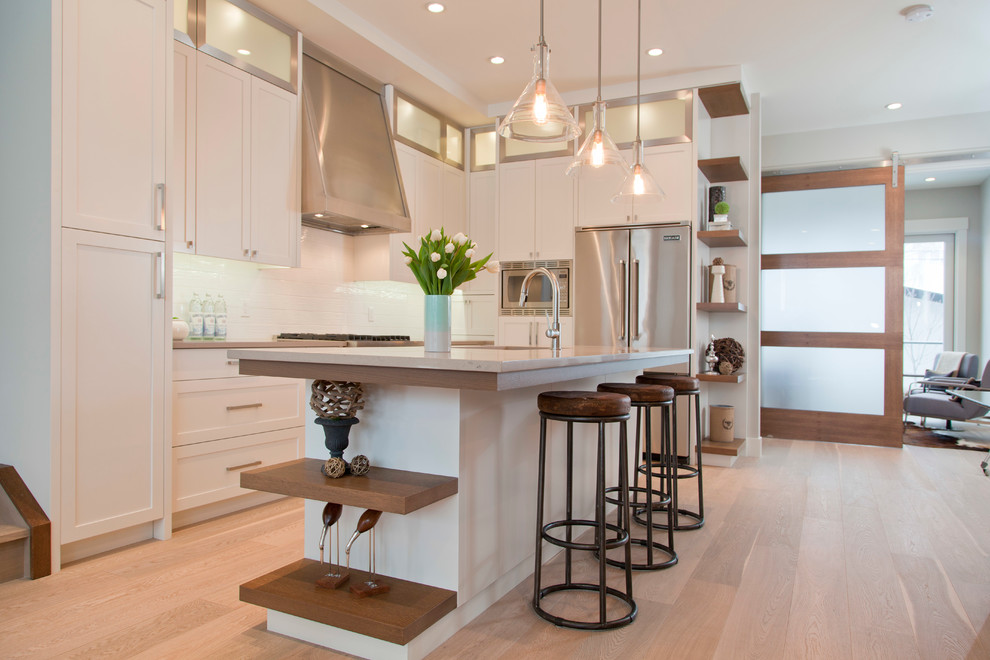 Diseño de cocina clásica renovada de tamaño medio con puertas de armario blancas, salpicadero blanco, electrodomésticos de acero inoxidable, suelo de madera clara y una isla