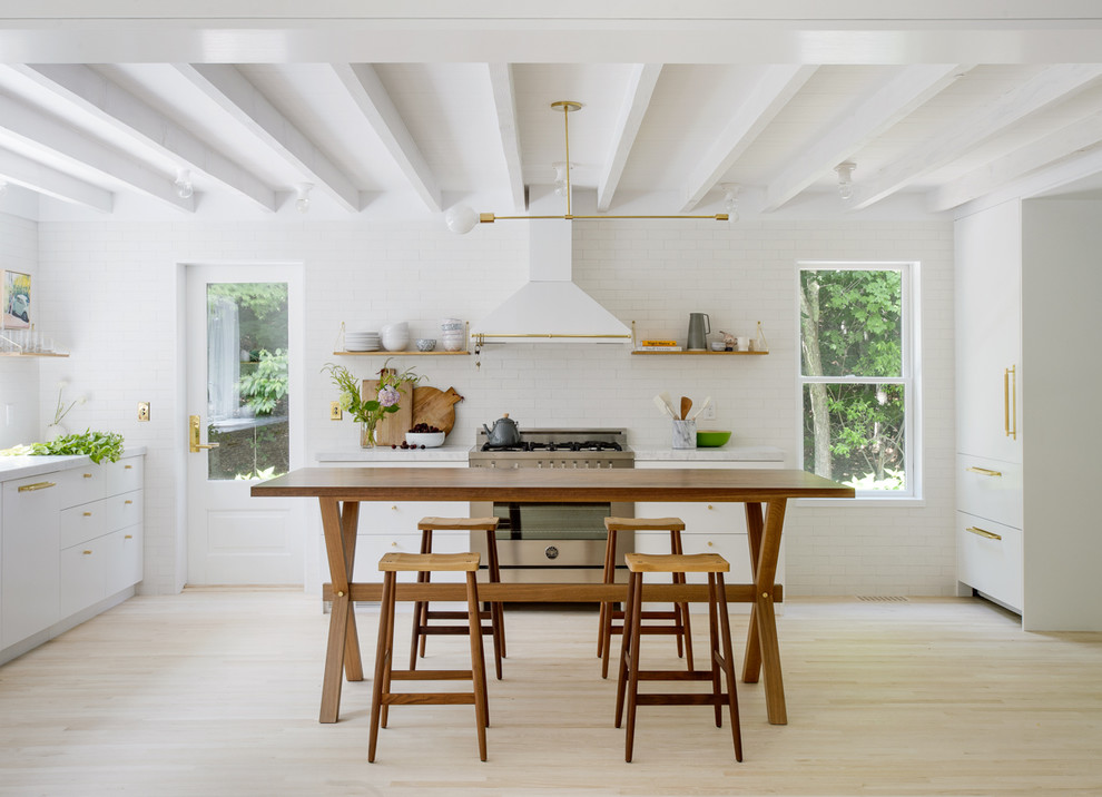 Bild på ett minimalistiskt vit vitt kök, med släta luckor, vita skåp, vitt stänkskydd, stänkskydd i tunnelbanekakel, rostfria vitvaror, ljust trägolv och en köksö