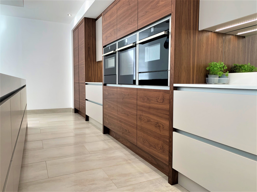 Large classic single-wall kitchen in Other with an integrated sink, flat-panel cabinets, medium wood cabinets, composite countertops, glass sheet splashback, black appliances, porcelain flooring, an island and white worktops.