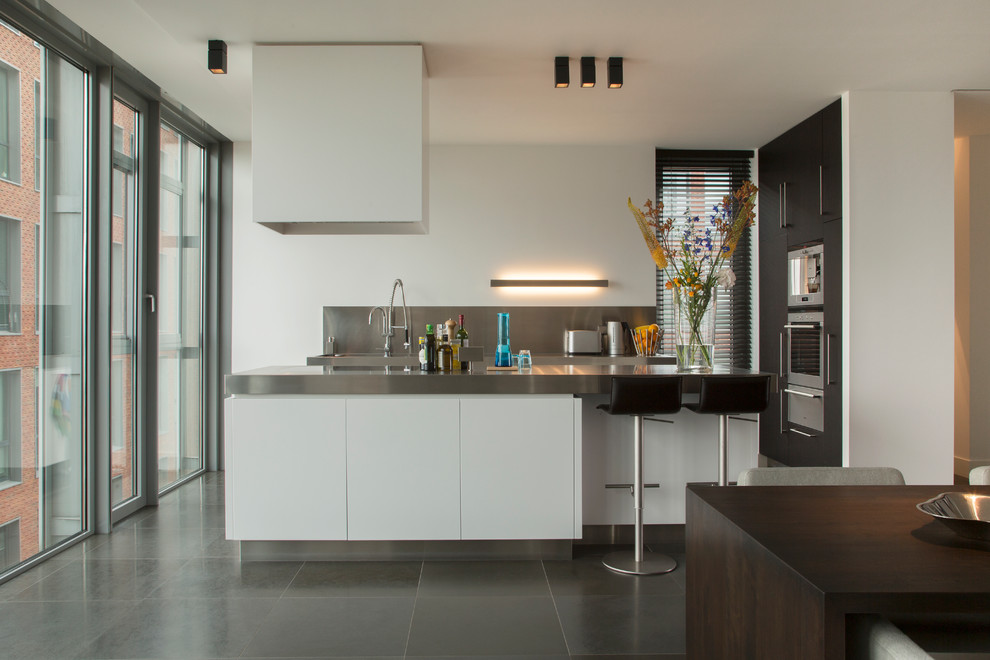 Inredning av ett modernt kök och matrum, med släta luckor, vita skåp, bänkskiva i rostfritt stål, stänkskydd med metallisk yta, rostfria vitvaror, en köksö och grått golv