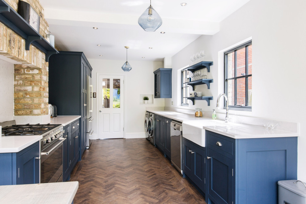 This is an example of a medium sized traditional galley kitchen/diner in Essex with a belfast sink, shaker cabinets, blue cabinets, quartz worktops, white splashback, brick splashback, stainless steel appliances, medium hardwood flooring, no island, brown floors and white worktops.