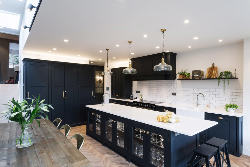 Design ideas for a large industrial galley kitchen/diner in London with a belfast sink, glass-front cabinets, blue cabinets, granite worktops, white splashback, ceramic splashback, black appliances, an island and white worktops.