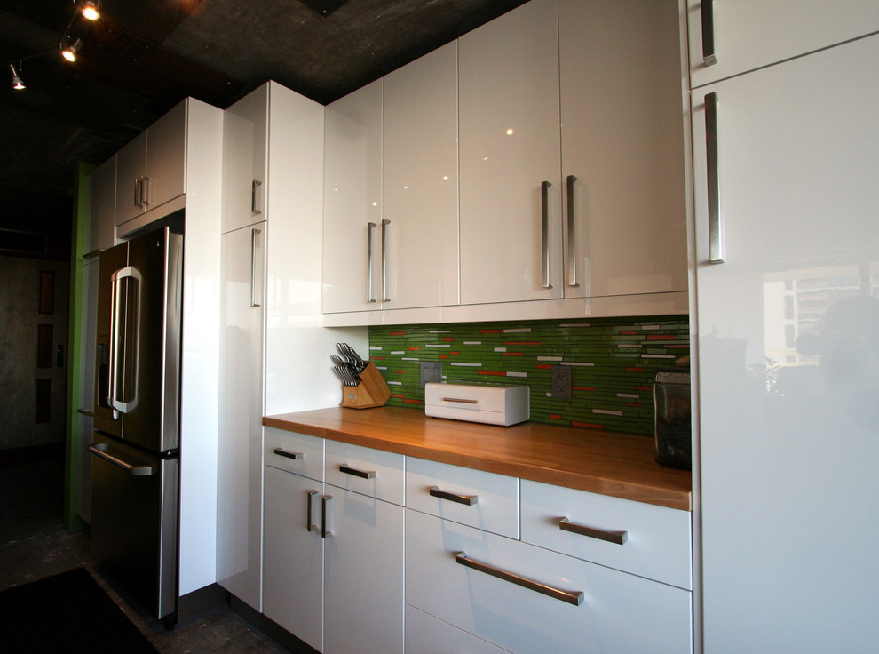 Small contemporary galley open plan kitchen in Other with a built-in sink, flat-panel cabinets, white cabinets, wood worktops, green splashback, glass tiled splashback, stainless steel appliances, concrete flooring and a breakfast bar.