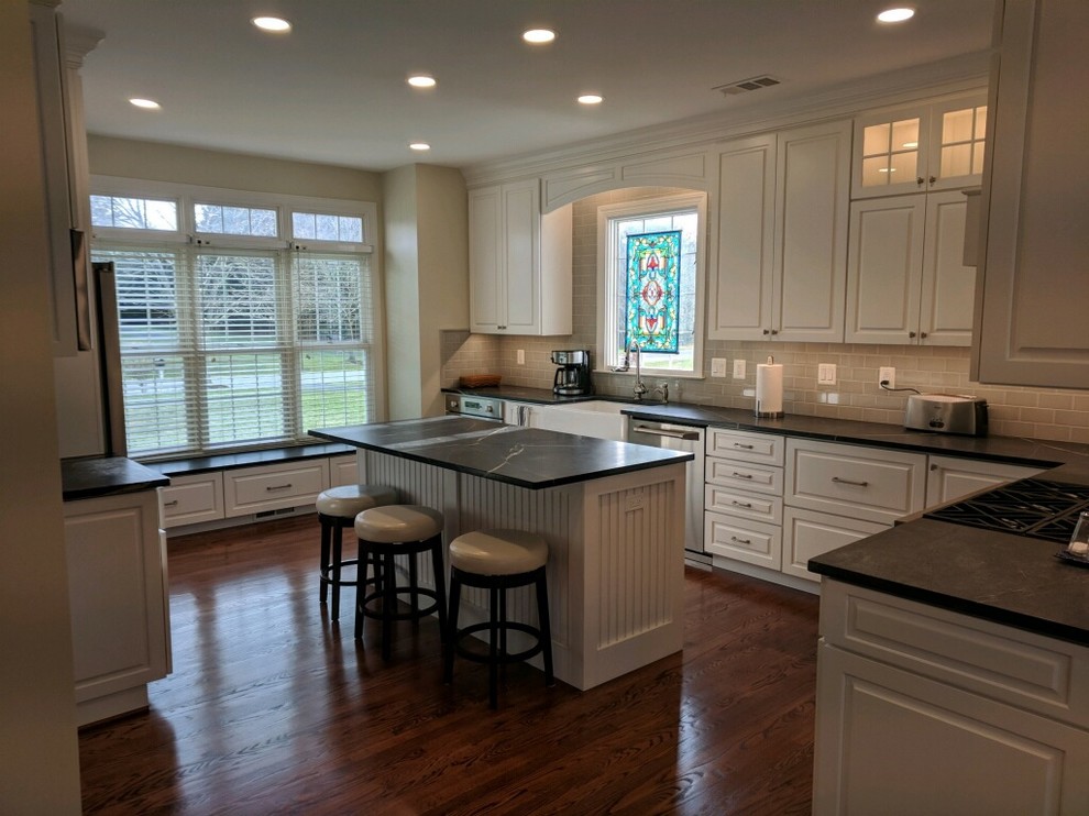 Large transitional l-shaped dark wood floor and brown floor eat-in kitchen photo in Baltimore with a farmhouse sink, raised-panel cabinets, white cabinets, soapstone countertops, gray backsplash, subway tile backsplash, stainless steel appliances and an island