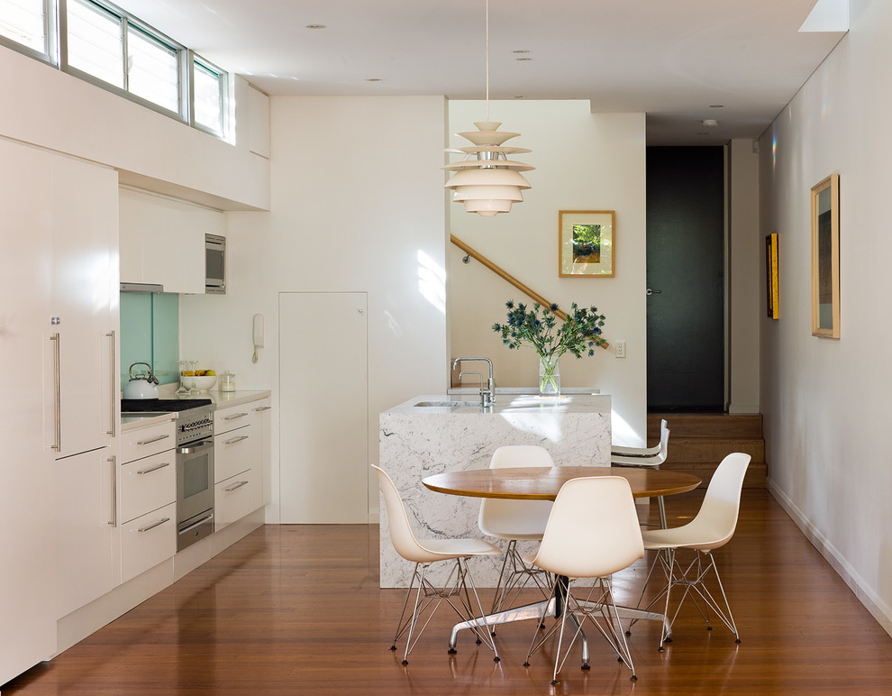 Modern inredning av ett kök, med en undermonterad diskho, vita skåp, marmorbänkskiva, glaspanel som stänkskydd, rostfria vitvaror, en köksö, brunt golv, släta luckor, blått stänkskydd och mörkt trägolv