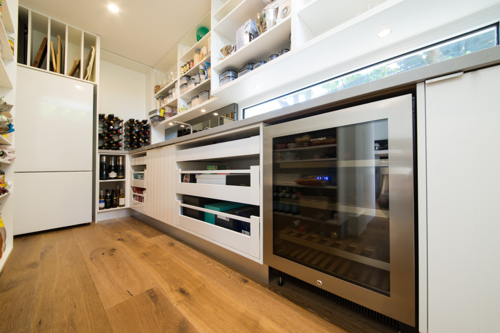 Design ideas for an expansive beach style galley kitchen pantry in Melbourne with a submerged sink, open cabinets, white cabinets, engineered stone countertops, metallic splashback, window splashback, stainless steel appliances, medium hardwood flooring, no island and brown floors.