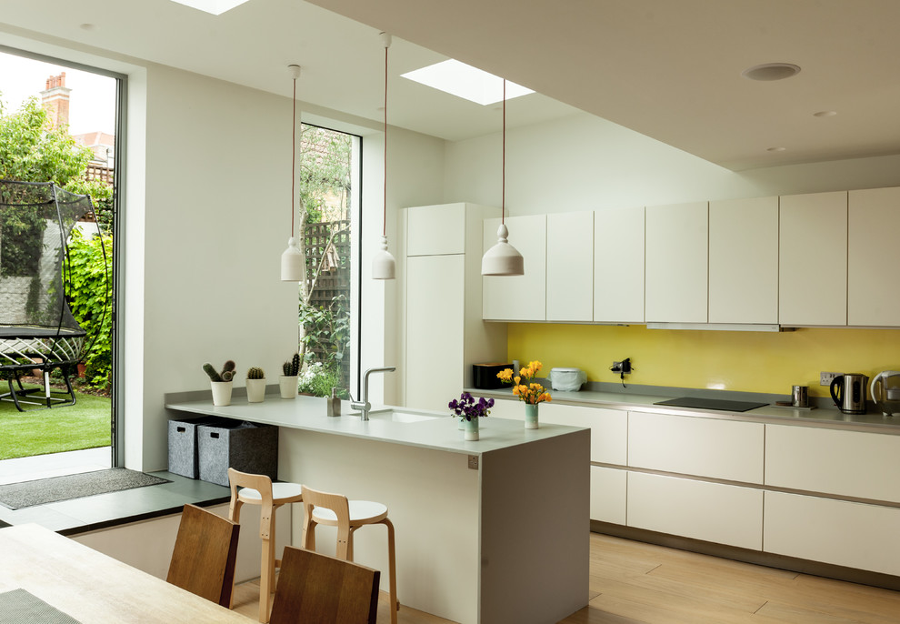 Contemporary galley kitchen in London with a submerged sink, flat-panel cabinets, white cabinets, yellow splashback, medium hardwood flooring, an island, brown floors and grey worktops.