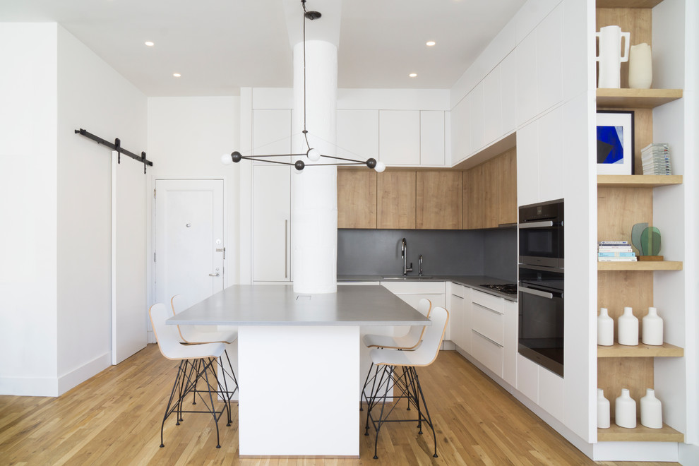 Example of a mid-sized trendy l-shaped medium tone wood floor and brown floor open concept kitchen design in New York with a drop-in sink, flat-panel cabinets, white cabinets, gray backsplash, black appliances, a peninsula, gray countertops, concrete countertops and cement tile backsplash