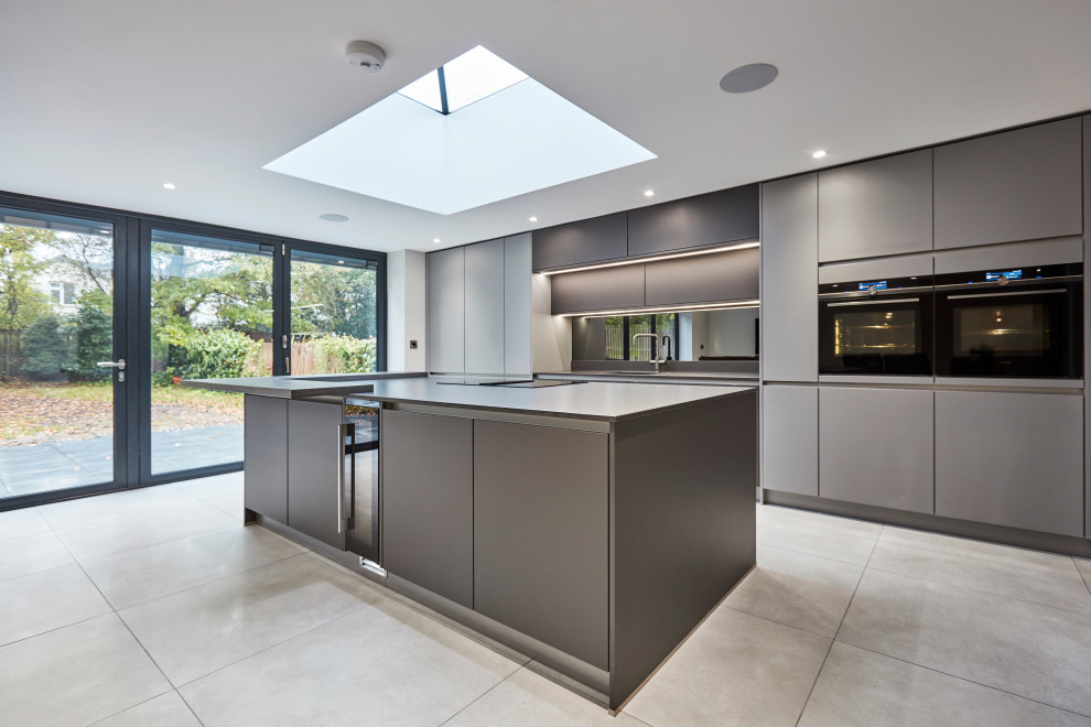 Photo of a medium sized contemporary single-wall open plan kitchen in Other with an integrated sink, flat-panel cabinets, grey cabinets, quartz worktops, metallic splashback, mirror splashback, black appliances, porcelain flooring, an island, grey floors, grey worktops and a vaulted ceiling.