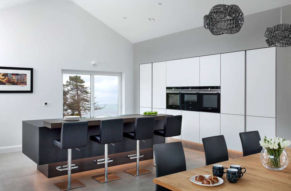 Идея дизайна: угловая кухня в современном стиле с обеденным столом, плоскими фасадами, белыми фасадами, черной техникой, полом из цементной плитки, островом и серым полом